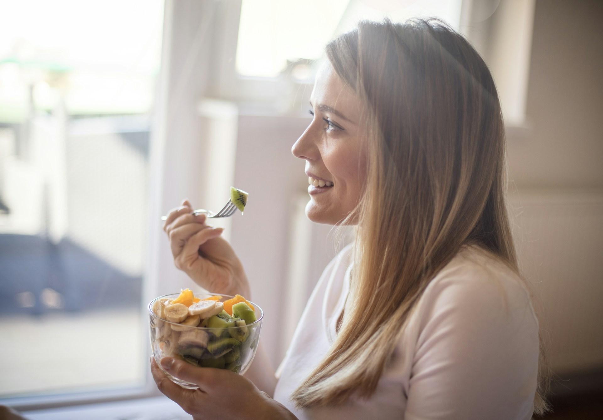 A woman stands next to a window and eats kiwi, banana, and mango out of a bowl. Sun shines through and reflects off of her blonde hair.