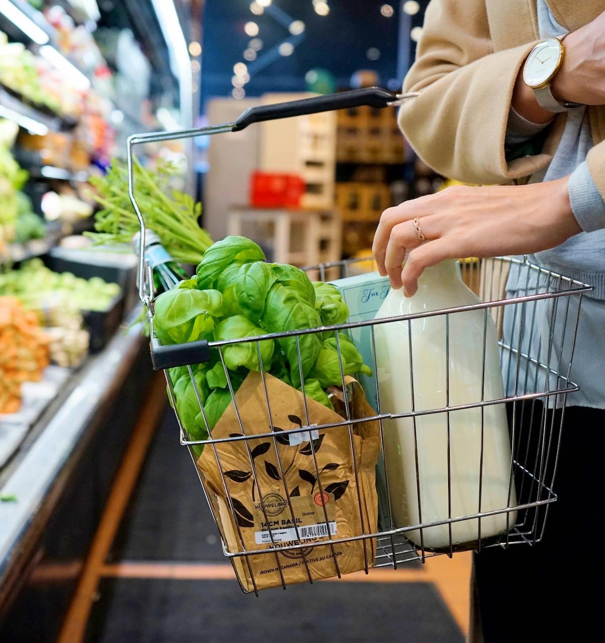 A well-dressed shopper adds a carton of milk to a silver grocery basket next to a large bunch of basil and other produce.